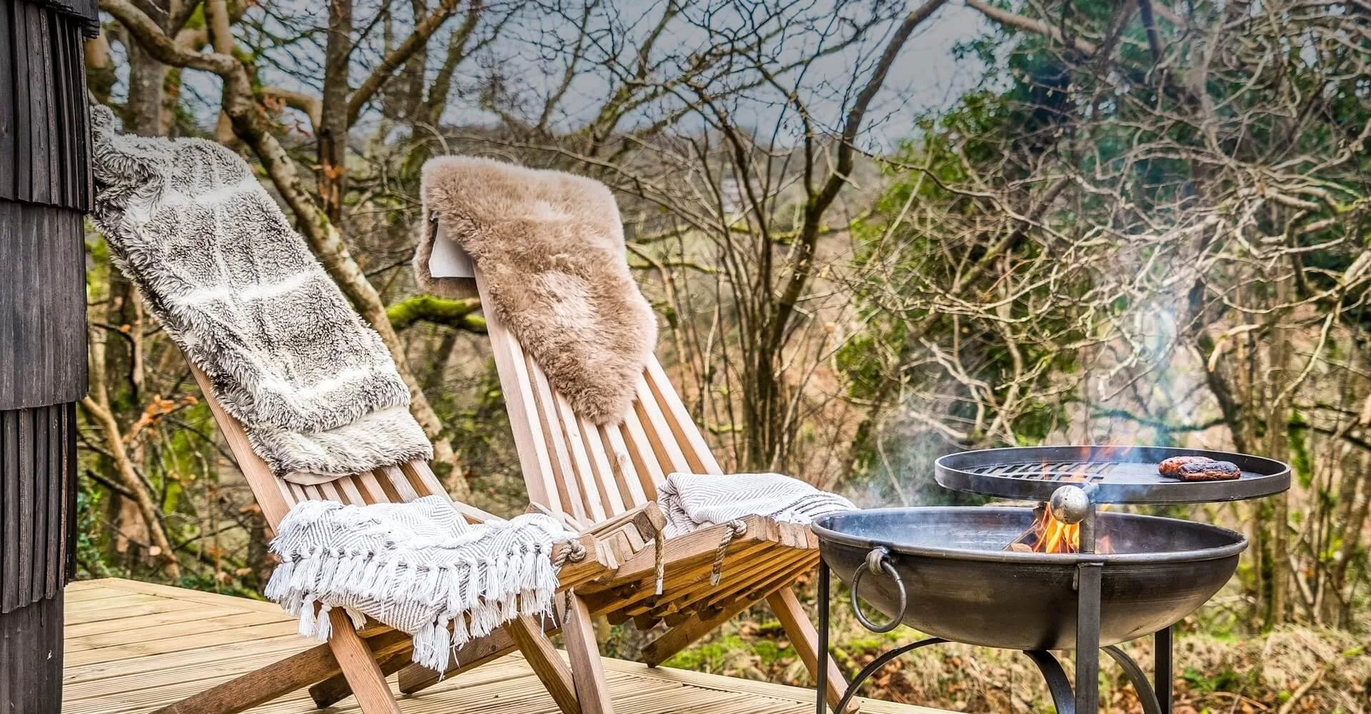 Two wooden lounge chairs with soft blankets draped over the top sit on a wooden deck next to a fire pit grill. One blanket is gray and fluffy, the other is light brown fur. In the background, there are trees with no leaves, indicating a fall or winte