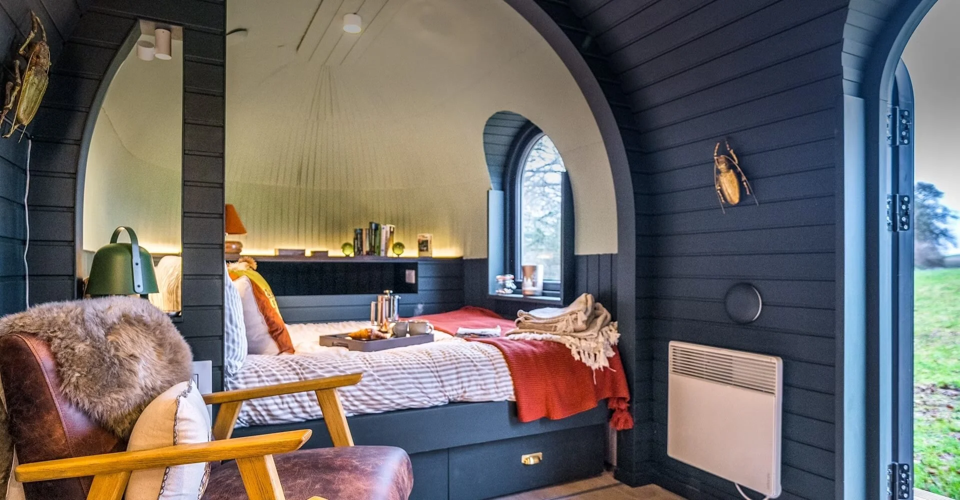 Interior of a cozy bedroom with dark wooden walls, an arched window, and a bed with striped bedding, pillows, and blankets. There is a wooden armchair with cushions and a faux fur throw. On the wall, there are decorative beetle sculptures.