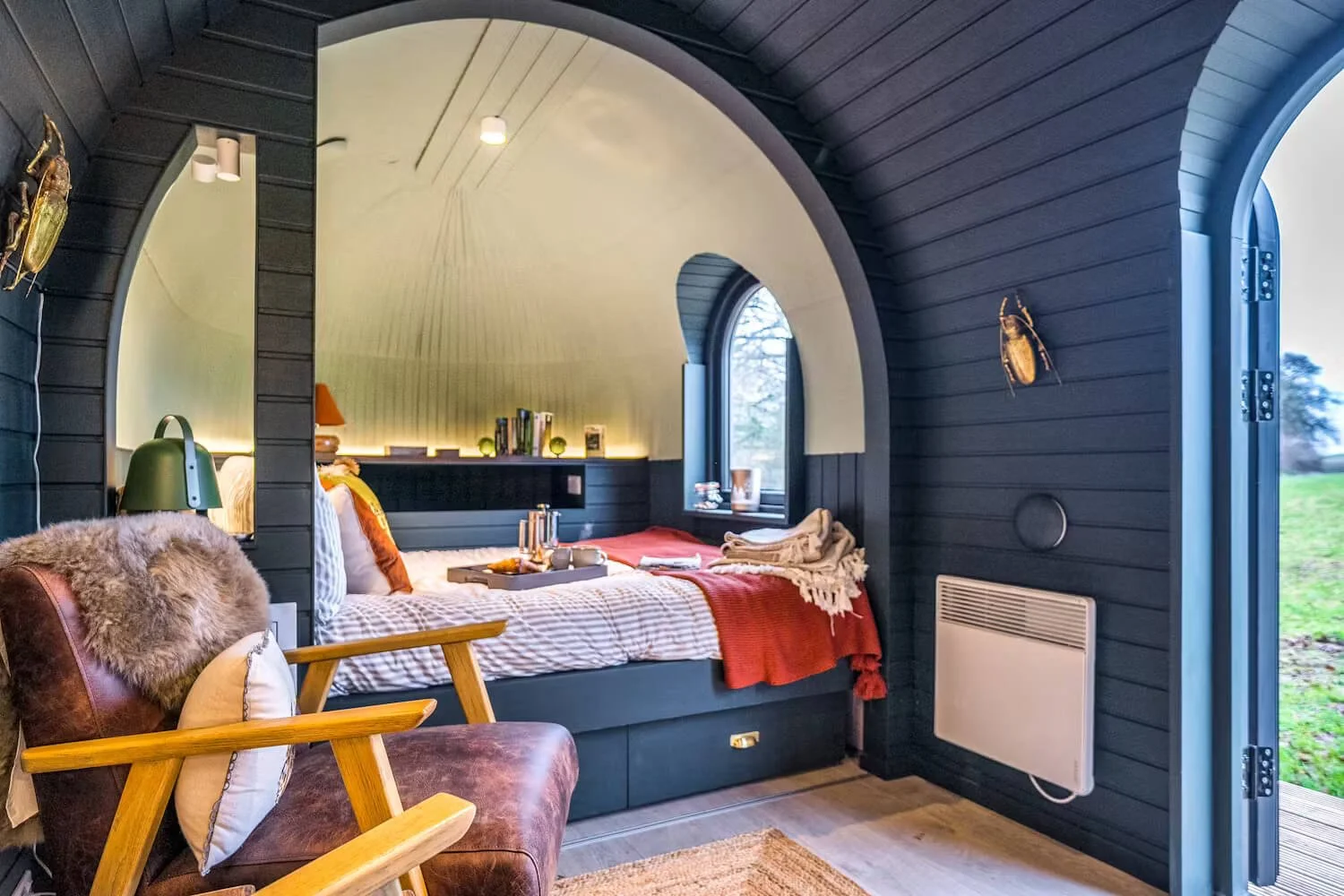Cozy bedroom with dark wood walls, arched ceiling, and large window, furnished with a bed, armchair, and various decor items, overlooking a green outdoor landscape.