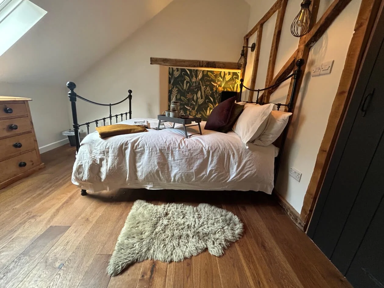 A bedroom with a white bed, decorative pillows, a tray with a jug and glasses, a wooden dresser, a cream shaggy rug, and decorative lighting fixtures, in a cozy, rustic style.