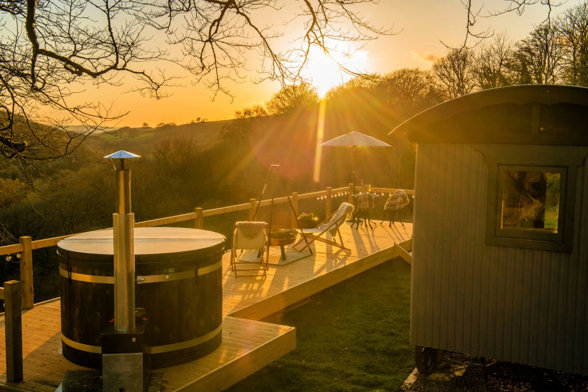 Sunset over a wooden deck with outdoor furniture, umbrella, hot tub, and a small wooden shed in a rural landscape with rolling hills and leafless trees.