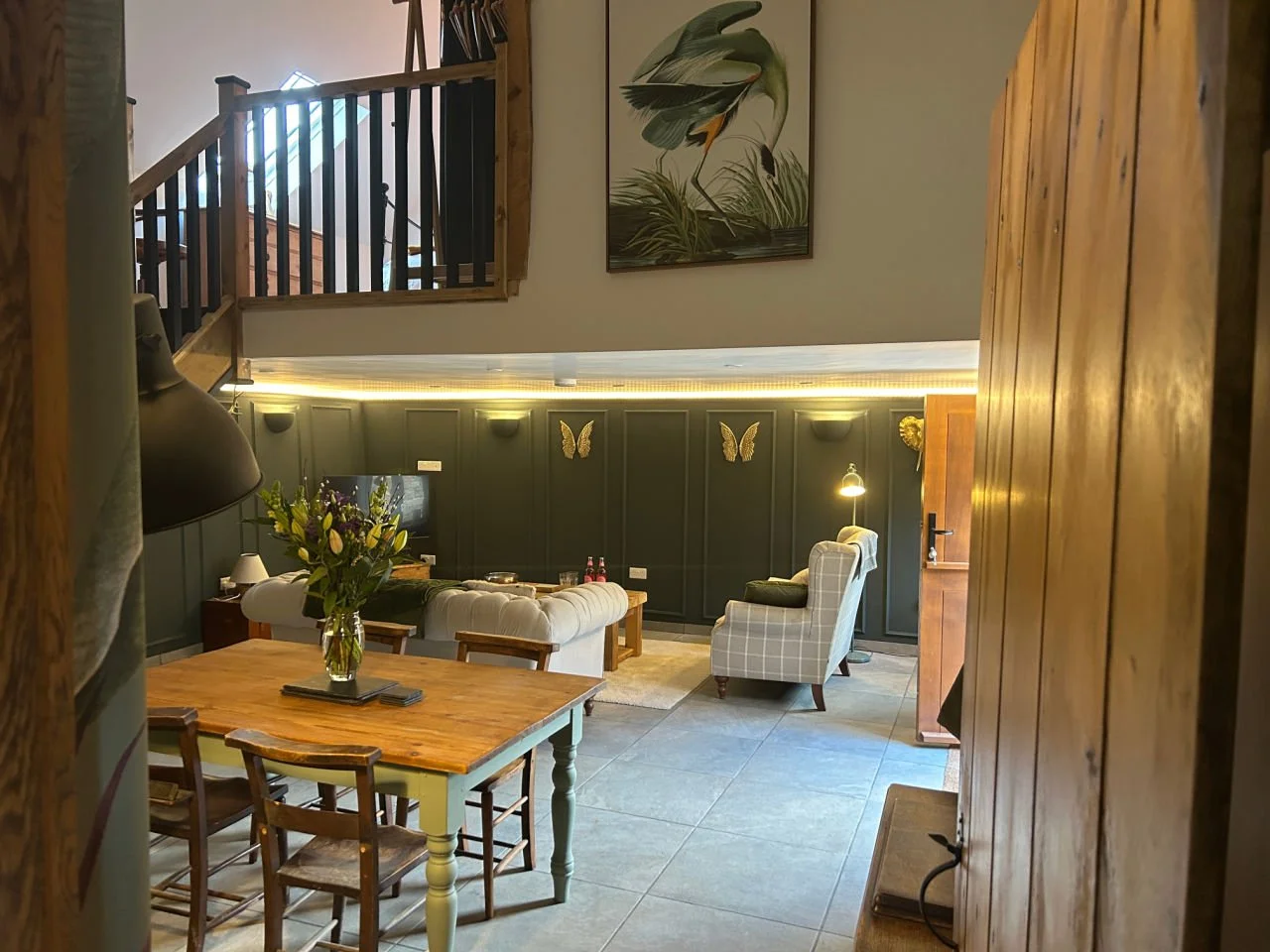Interior of a cozy living room and dining area with dark green paneled walls, decorated with butterfly ornaments, a floral painting, and a variety of seating including a beige sofa and checkered armchair, illuminated by wall and floor lamps.