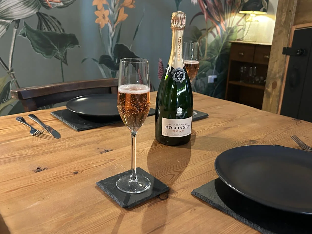 A wooden dining table set for a meal with a bottle of Bollinger Rose champagne and two glasses of champagne, dark plates, cutlery, and a slate placemat. The background has floral wallpaper and a wooden cabinet with glasses.