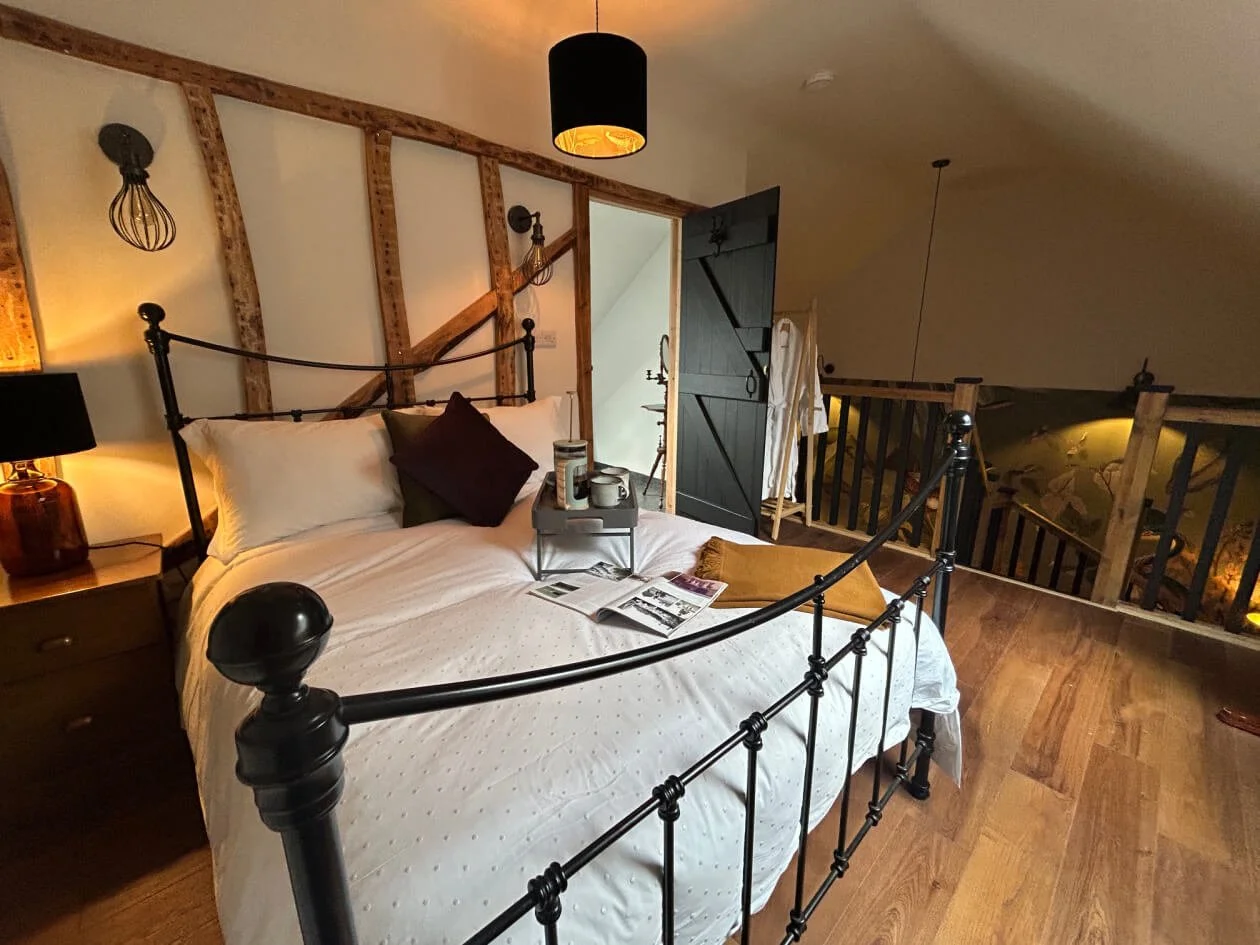 Cozy bedroom with a brass bed, white bedding, and dark pillows. Wooden wall paneling behind the bed with industrial-style wall lamps. Nightstand with a lamp and decor items. Open door reveals a bathroom or closet area. Loft space with a wooden railin