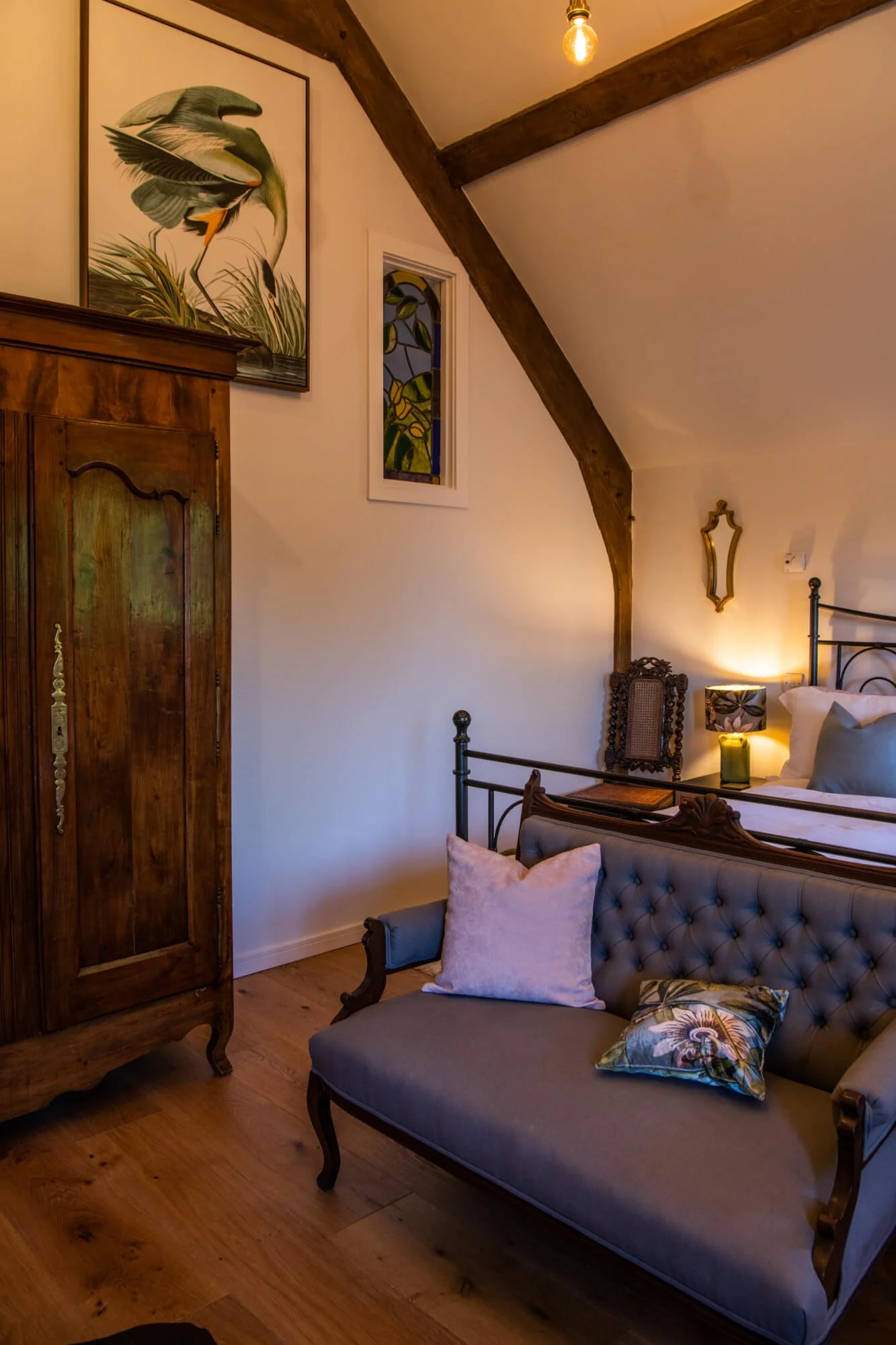 A cozy bedroom corner with a vintage armchair with pillows, a wooden wardrobe, a bed with pillows, a bedside table with a lamp, a small framed mirror, a stained glass window, and a wall art of a large bird and foliage, in a room with wood accents and