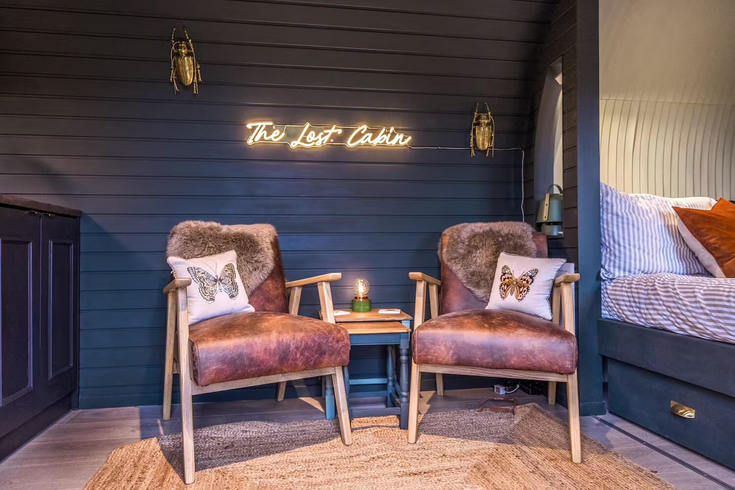 A cozy corner in a cabin with two brown leather armchairs, each with a butterfly pillow, a wooden side table between them with a small lamp, dark wood wall with a neon sign reading 'The Lost Cabin', and part of a bed with striped bedding on the right
