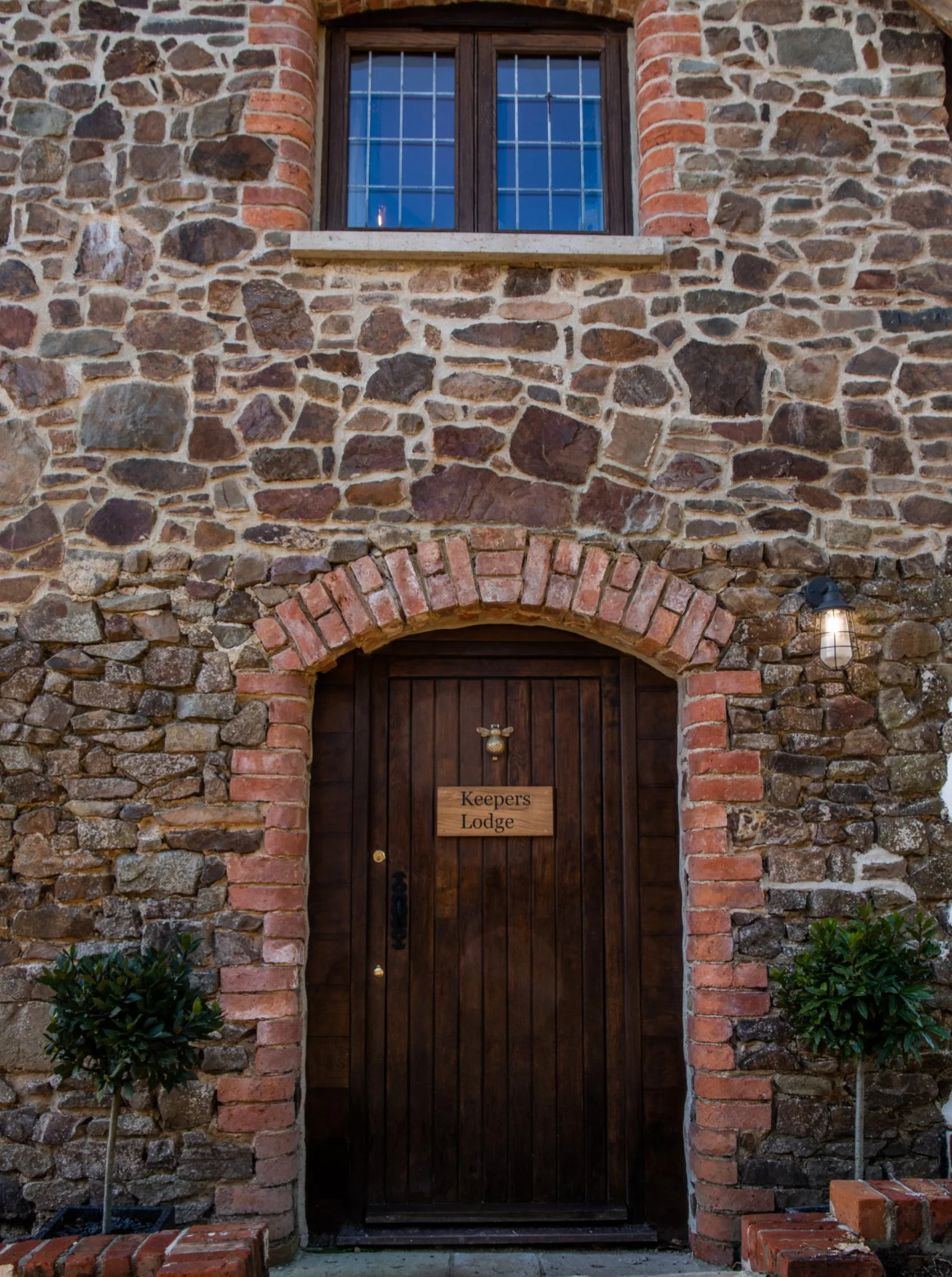 A rustic stone building with a wooden door labeled 'Keepers Lodge' and two potted bushes outside.