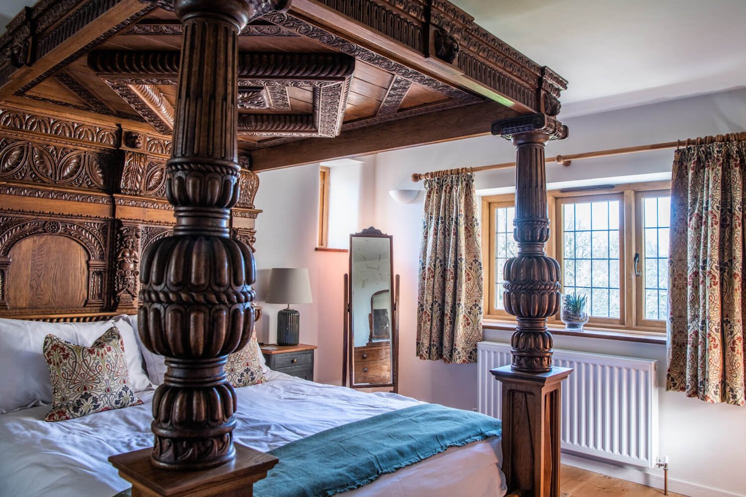A bedroom with a carved wooden four-poster bed, patterned curtains, a small table with a lamp, a full-length mirror, and a window with a view of trees outside.