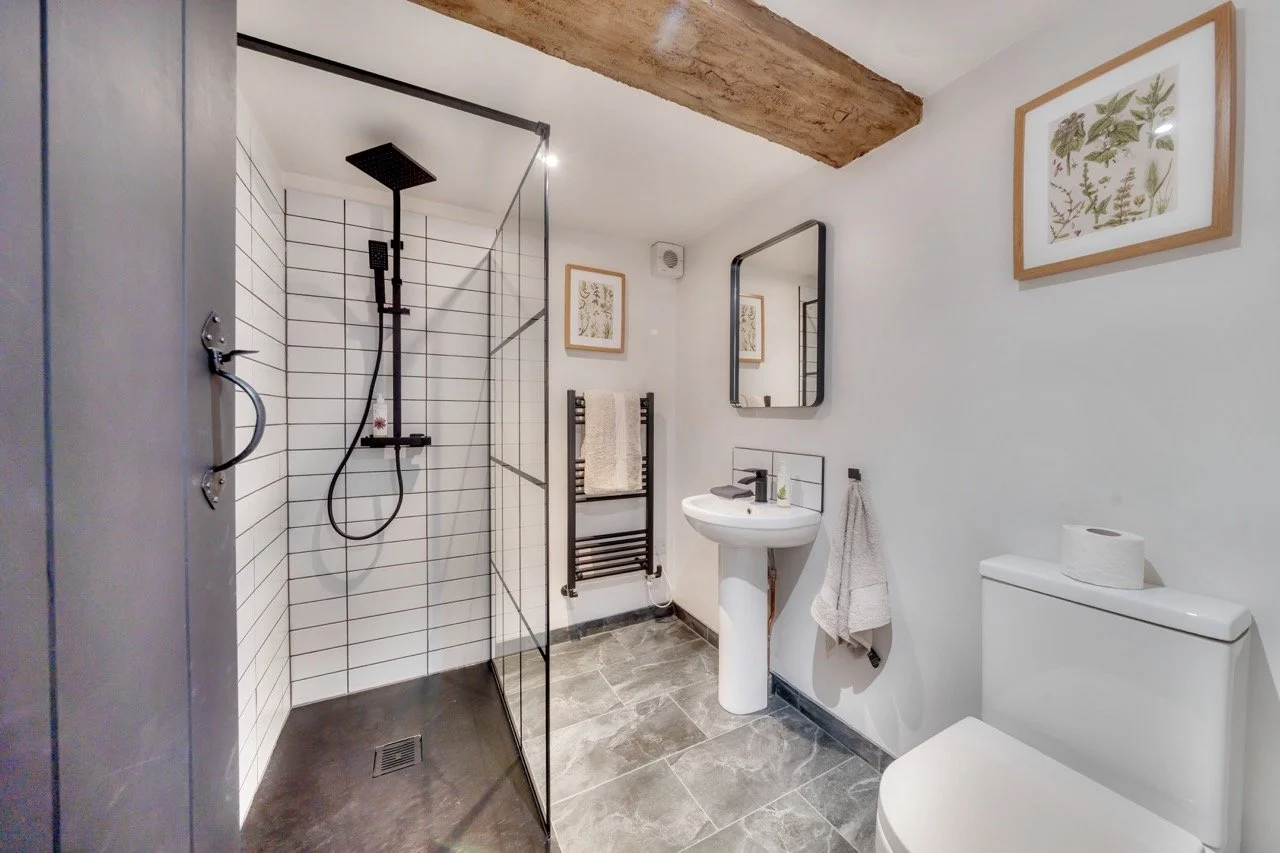 Modern bathroom with walk-in shower, white tiles, black fixtures, small sink with black faucet, mirror, framed botanical artwork, gray flooring, towel rack with towels, toilet, and wooden ceiling beam.