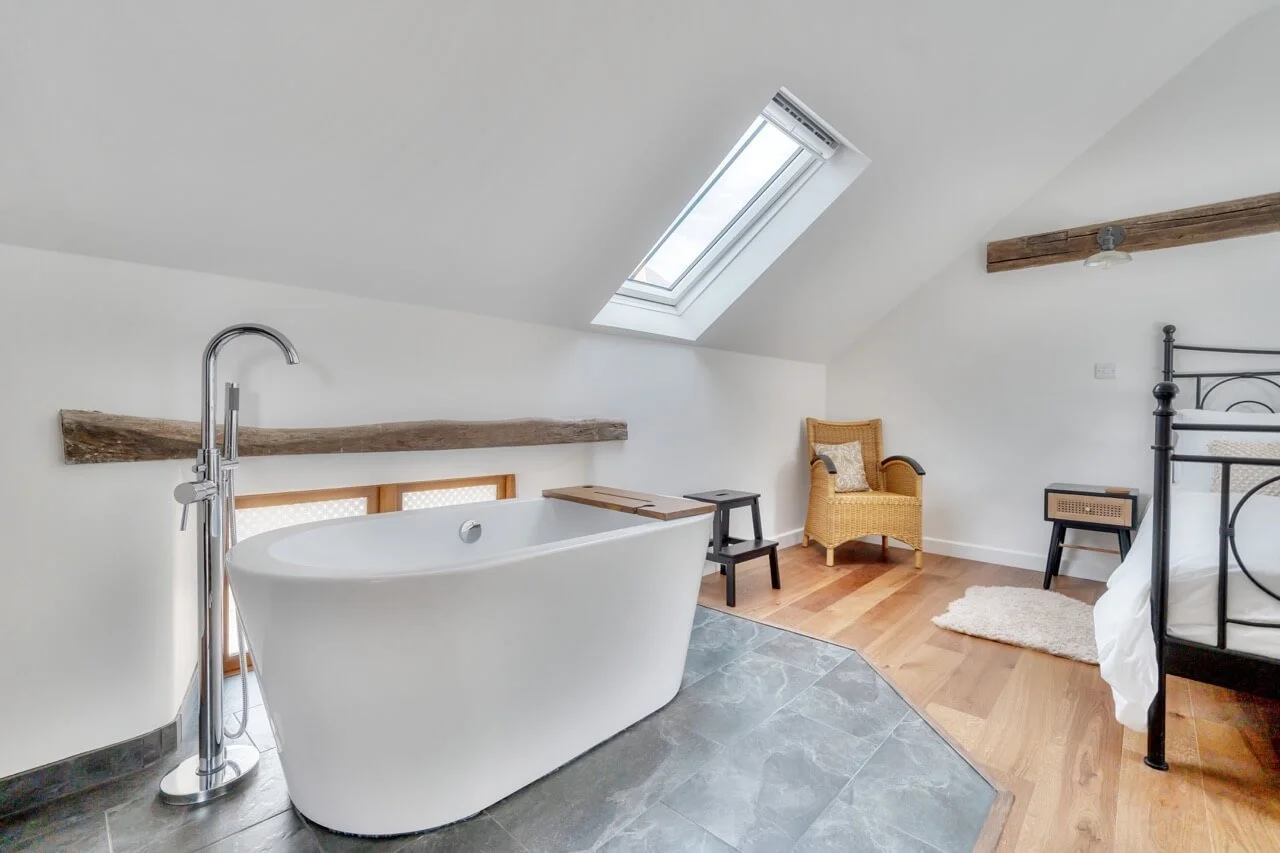 A cozy attic bathroom with a white freestanding bathtub, a skylight window, a black metal bed frame, a wicker armchair, small side tables, wooden beams, and wood flooring.