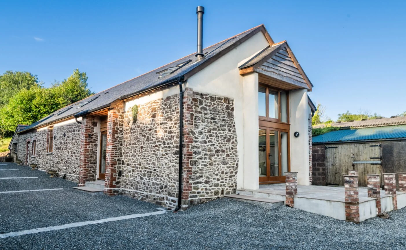 Tumbledown Barn a converted cow shed with a stone facade, wooden accents, and a metal chimney, featuring a glass front door, small steps, and parking spaces in front.