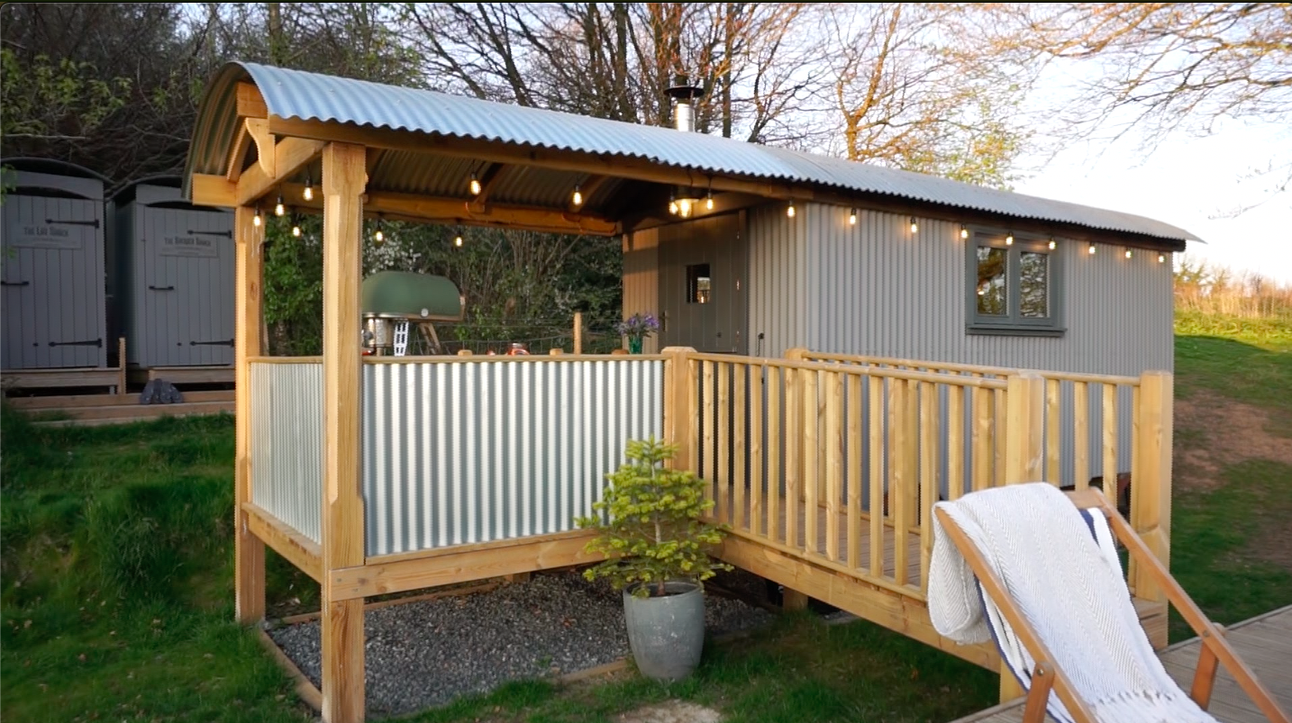 Small grey metal-clad building with a wooden porch and railing, string lights hanging from the roof, a potted plant in front, and an outdoor chair with a towel draped over it in the foreground.