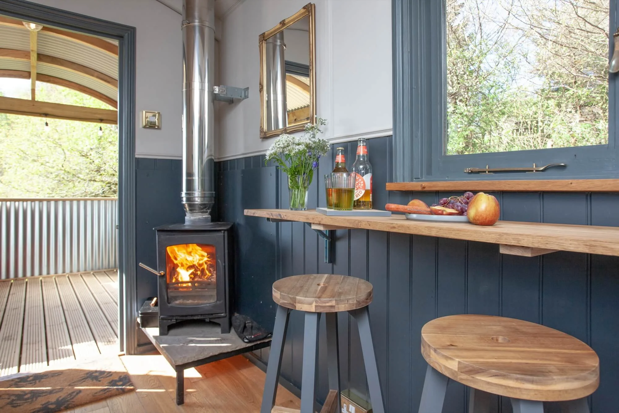 Interior of a cozy cabin with a wood-burning stove, a small wooden table, bar stools, a floating shelf with fruit and drinks, a window showing trees outside, and a door leading to an outdoor deck.