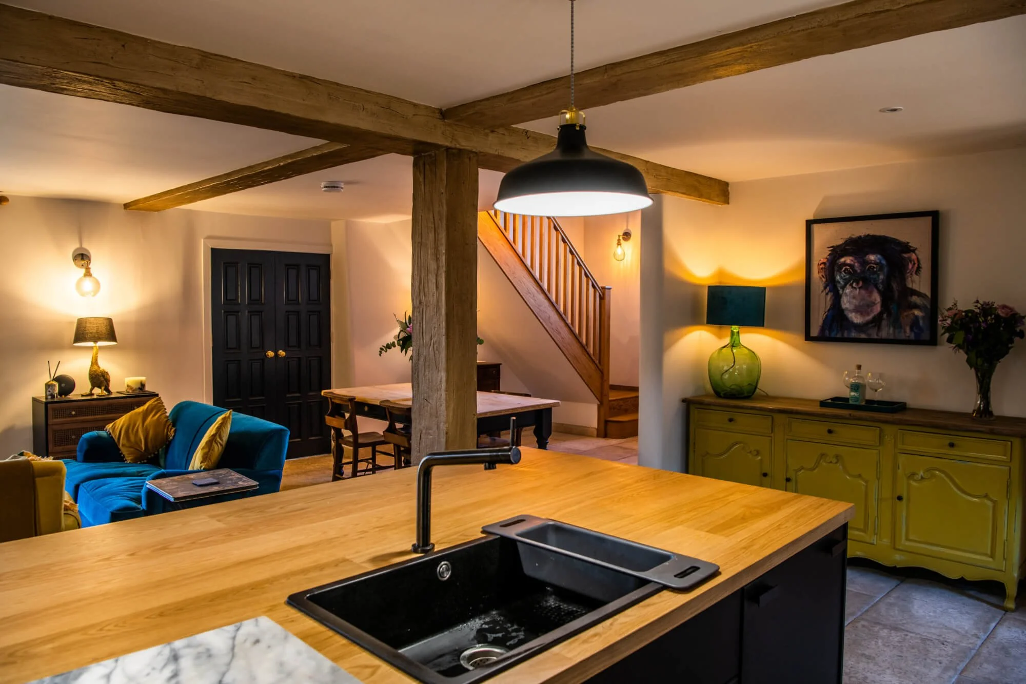 Living room with a blue sofa, yellow pillows, wooden dining table, modern black kitchen sink, green lampshade, large framed painting of a dog, staircase, and wooden beams on the ceiling.