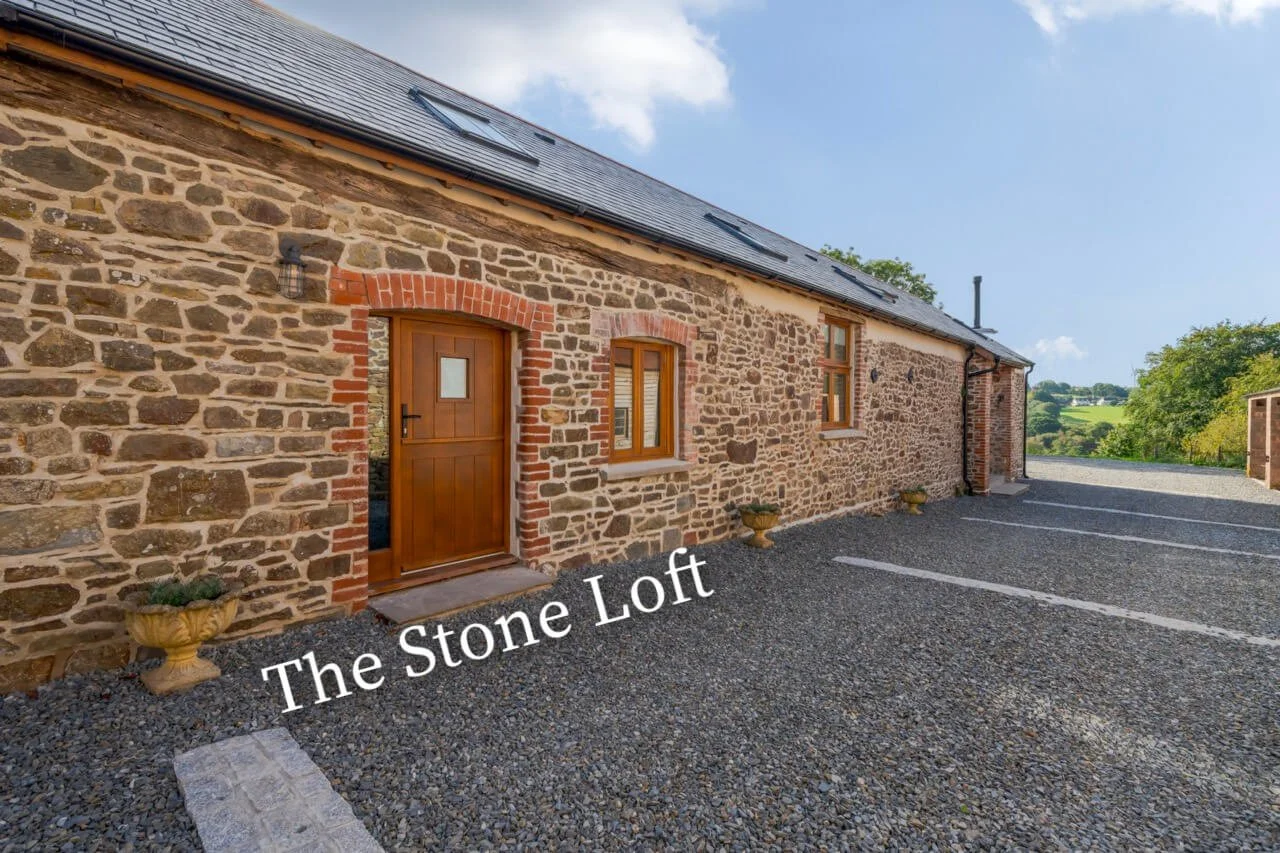 A stone brick building with a wooden door and two windows, surrounded by a gravel parking lot, with rolling green hills and blue sky in the background.