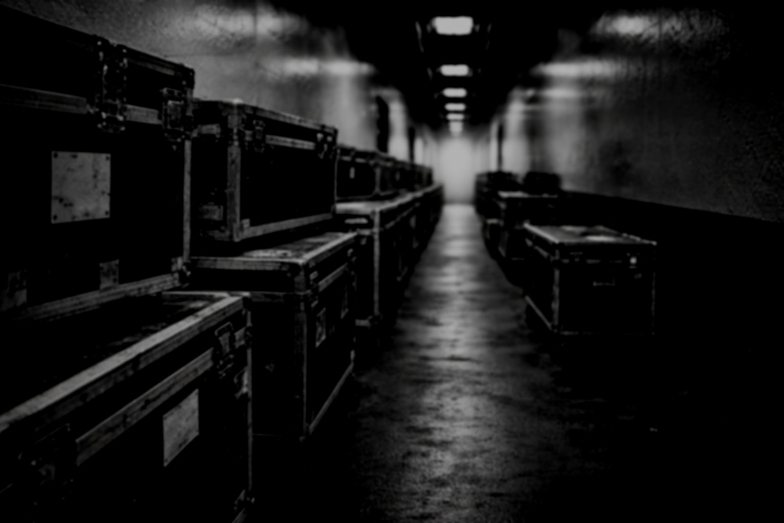A dark, narrow corridor with storage boxes on both sides, illuminated by overhead lights, leading to a bright exit at the end.