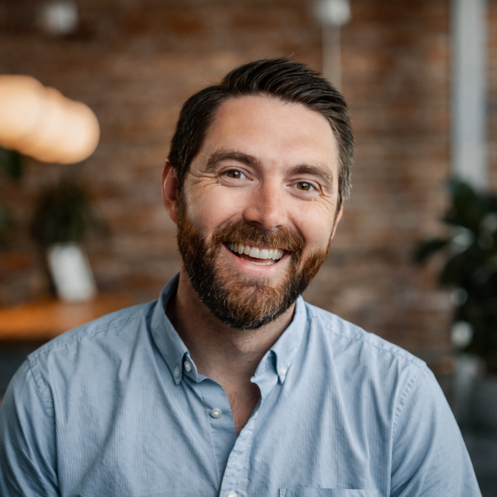 A man with a beard and short dark hair smiling at the camera, wearing a light blue button-up shirt, in a warmly lit room with brick wall and plants in the background.