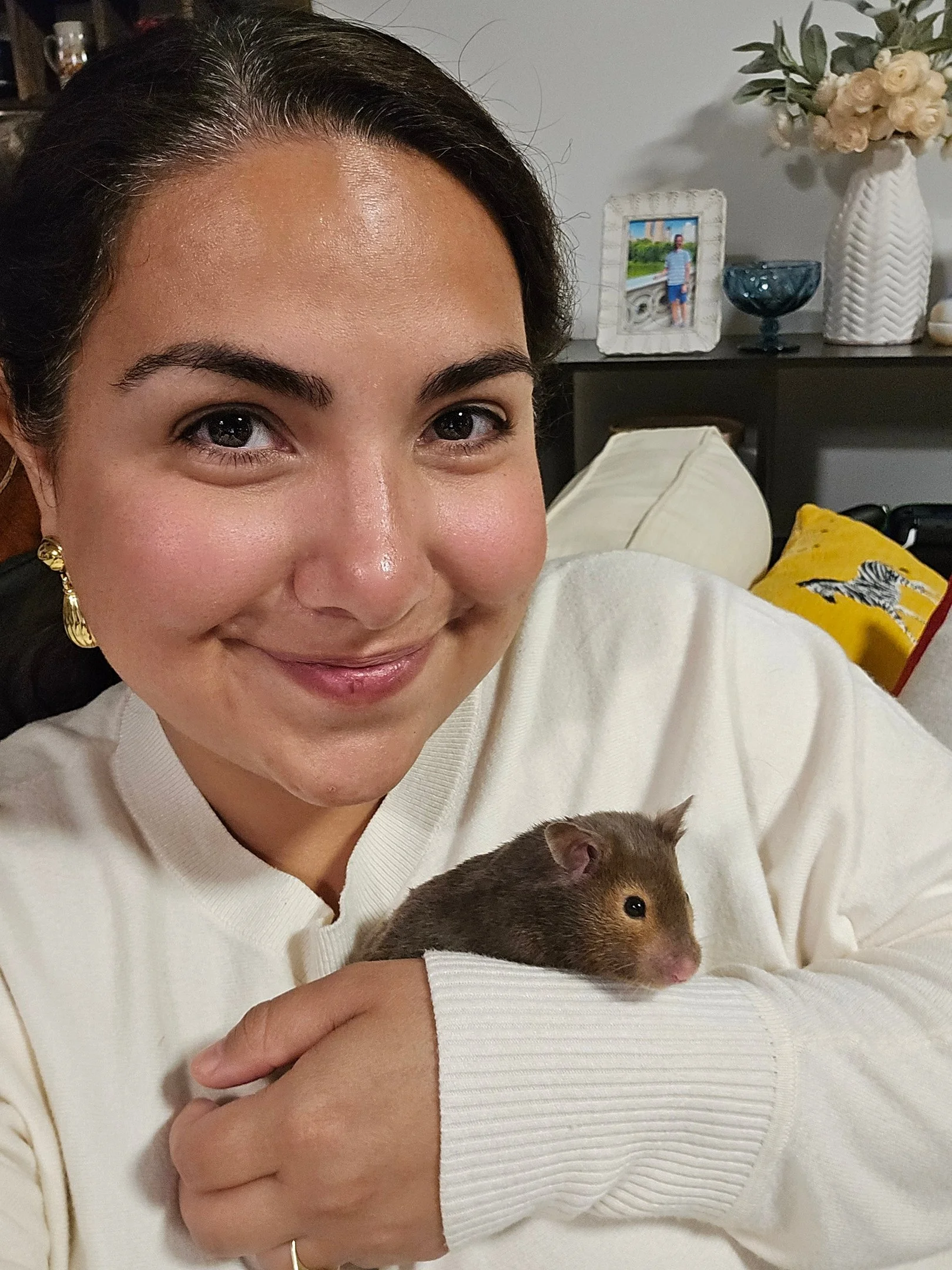 A woman smiling and holding a small hamster in her arm, sitting on a couch in a living room with decorative pillows and a shelf with picture frames, vases, and other decorative items in the background.