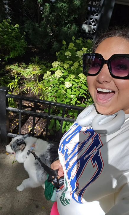 A woman wearing sunglasses and headphones taking a selfie with her dog in a garden surrounded by green plants and flowers.