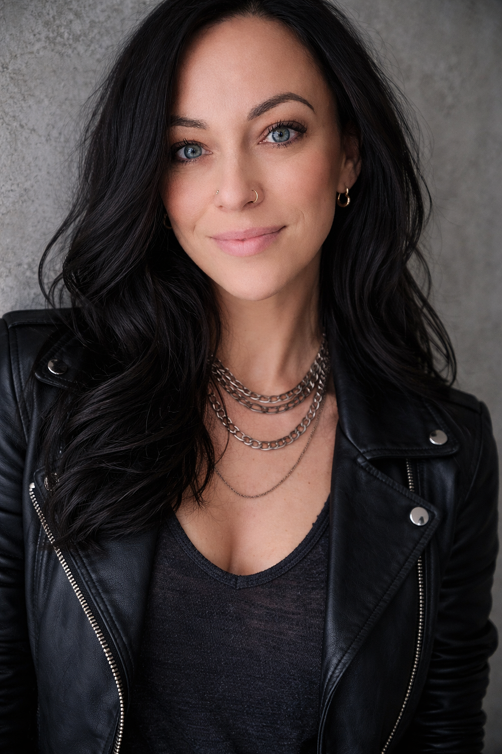 A woman with long dark wavy hair, striking blue eyes, wearing a black leather jacket, layered silver necklaces, and small hoop earrings, standing against a gray textured background.