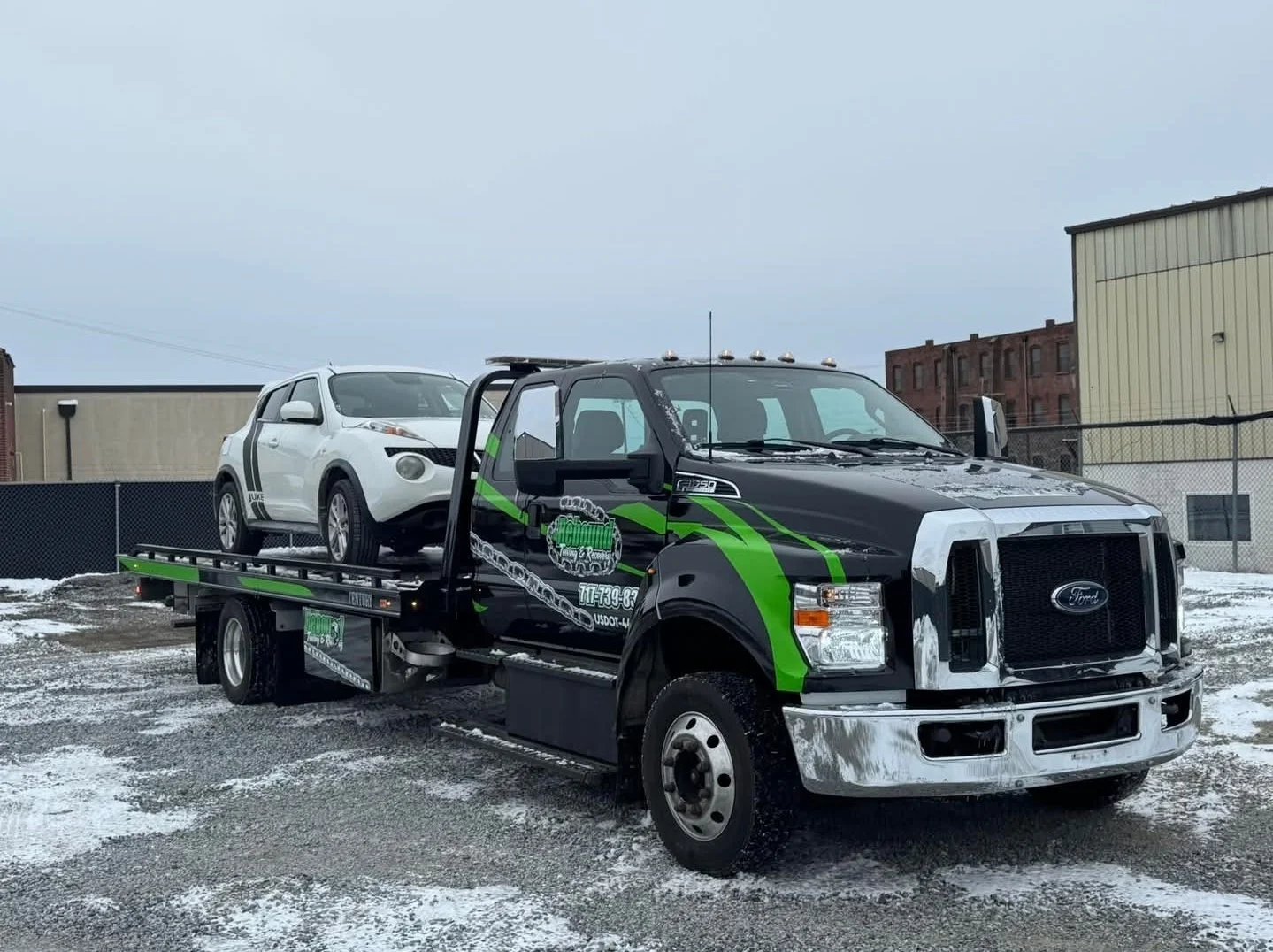 Black and green tow truck transporting a white SUV in an outdoor lot with snow-covered ground.