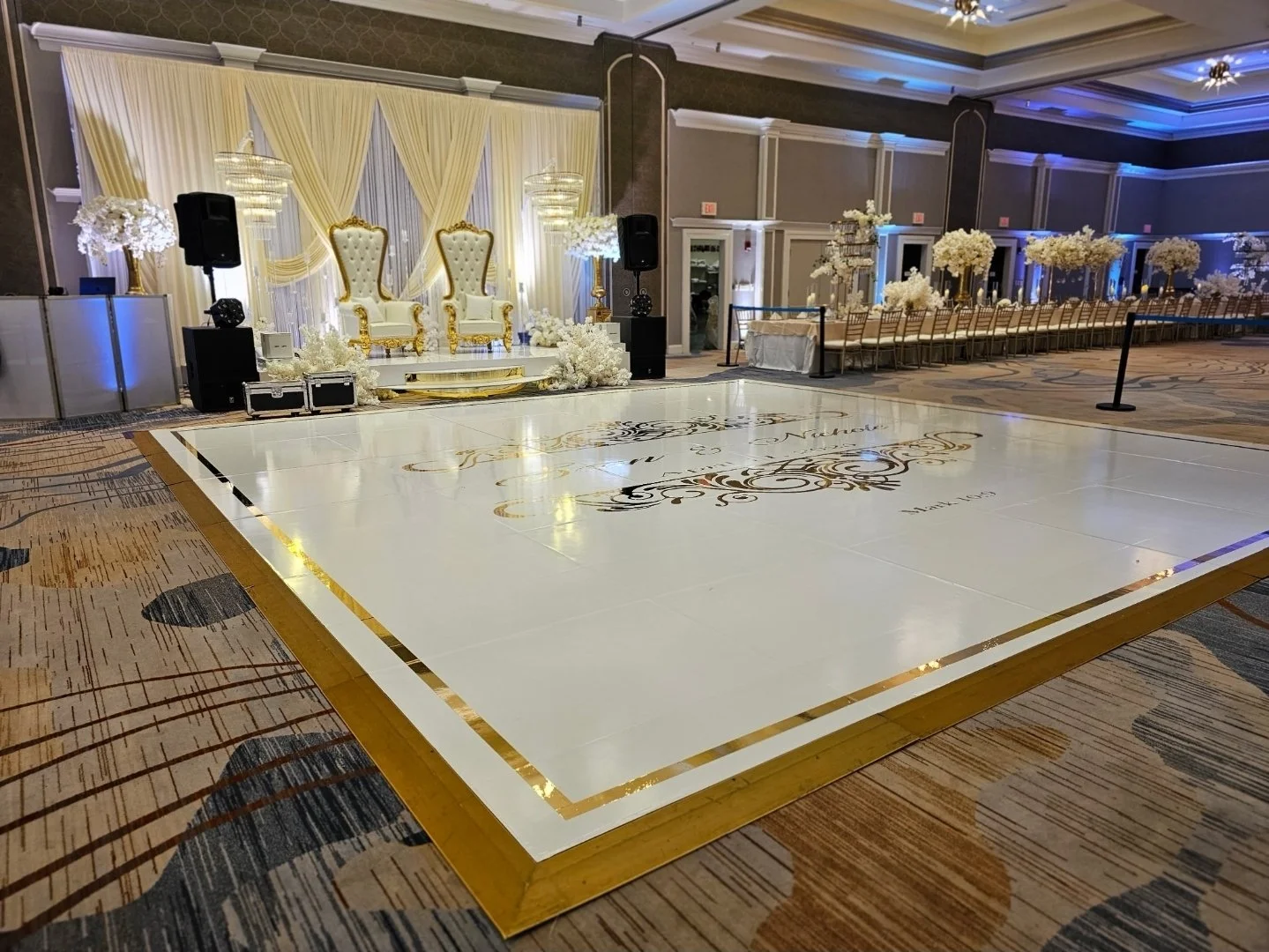 Elegant wedding or event setup with a decorated dance floor, ornate throne chairs on a stage, and tables lined with floral centerpieces in a large, decorated banquet hall.