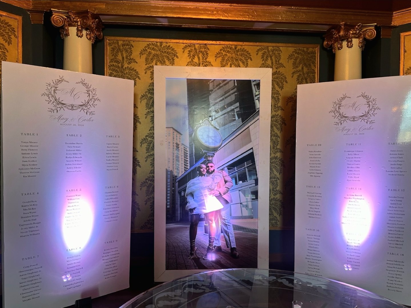 Large framed photograph of two people standing outside in a city with high-rise buildings, under a clear sky. One person is wearing a white coat and the other a light-colored suit, both looking at the camera. The display is inside a decorated room wi