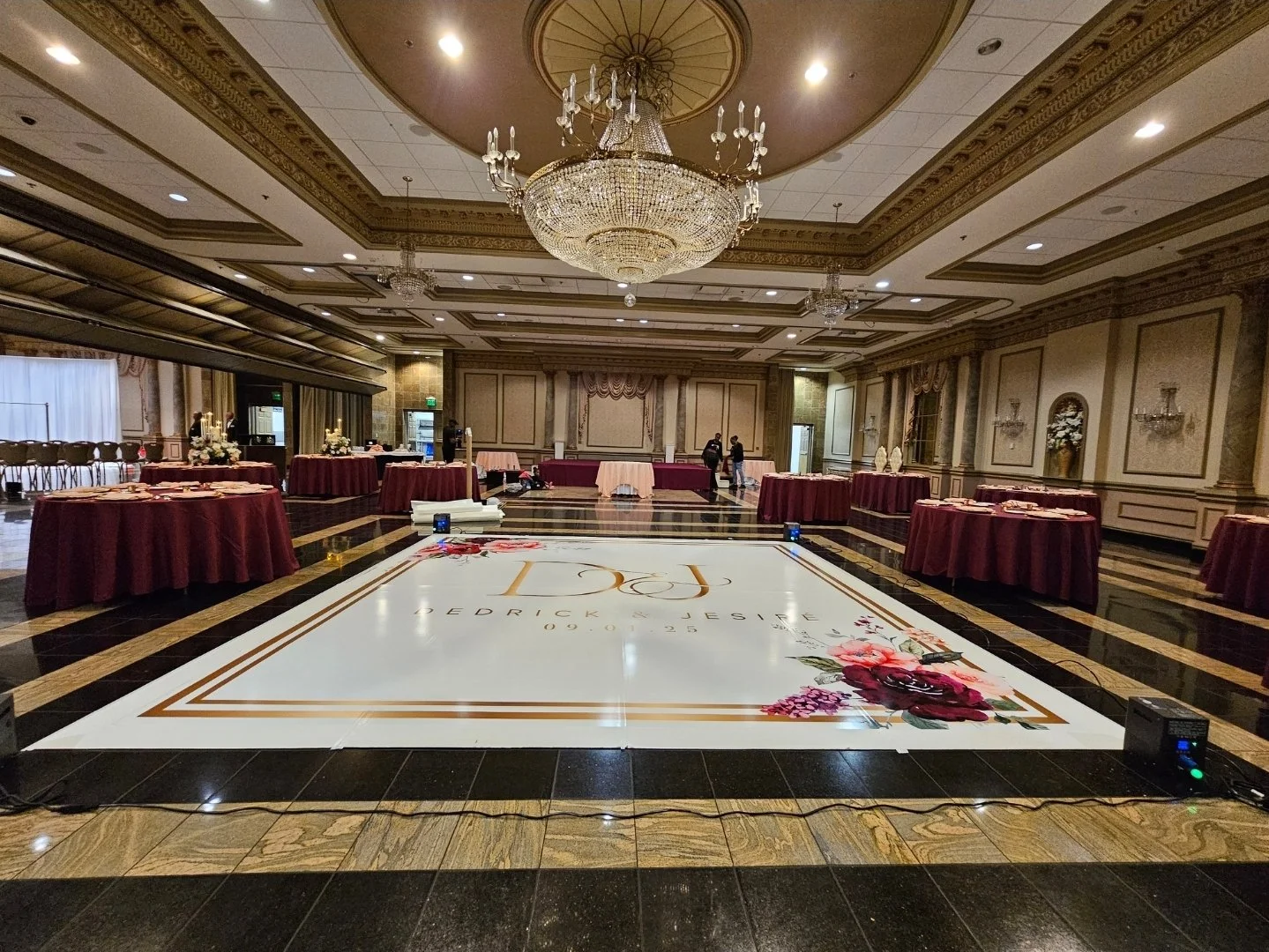 Elegant ballroom with a large chandelier, decorated tables with burgundy tablecloths, and a custom dance floor with floral designs and event details.