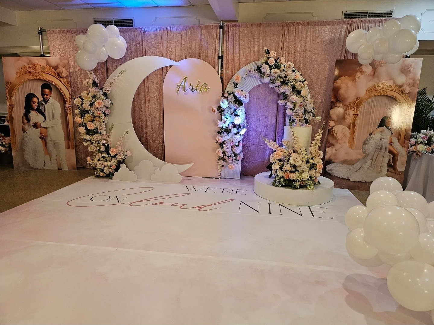 Wedding event space with a backdrop of pink curtains, floral arrangements, and large decorative letters spelling "Aria". Photos of a couple in wedding attire are displayed on either side. There are white balloons and clouds, with a floor projection t