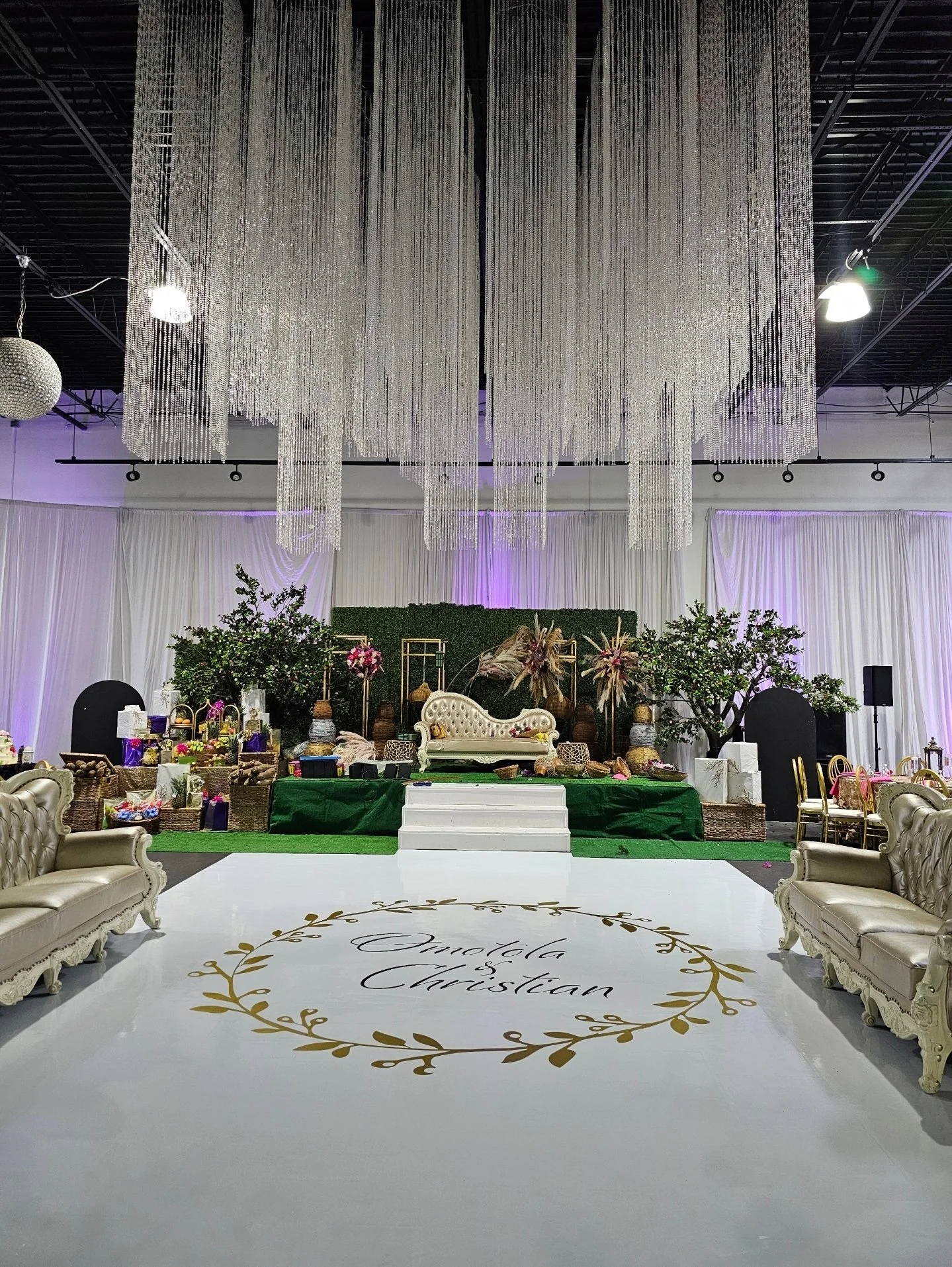 Indoor wedding setup with a white dance floor featuring a gold floral design and the names 'Onatola & Christian'. There is a vintage-style white sofa on a green stage, surrounded by various decorative items, flowers, and gifts. The ceiling is decorat