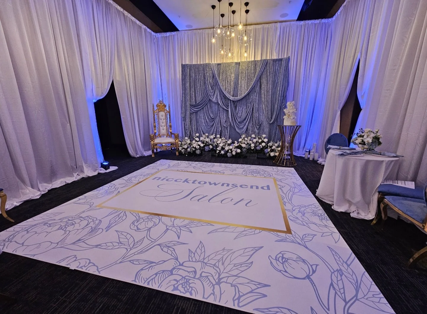 Elegant event stage with white drapes, blue lighting, a floral stage backdrop, a gold ornate chair, and floral arrangements on side tables, with a large white carpet featuring floral patterns and the text 'nicktownsend Salon'.