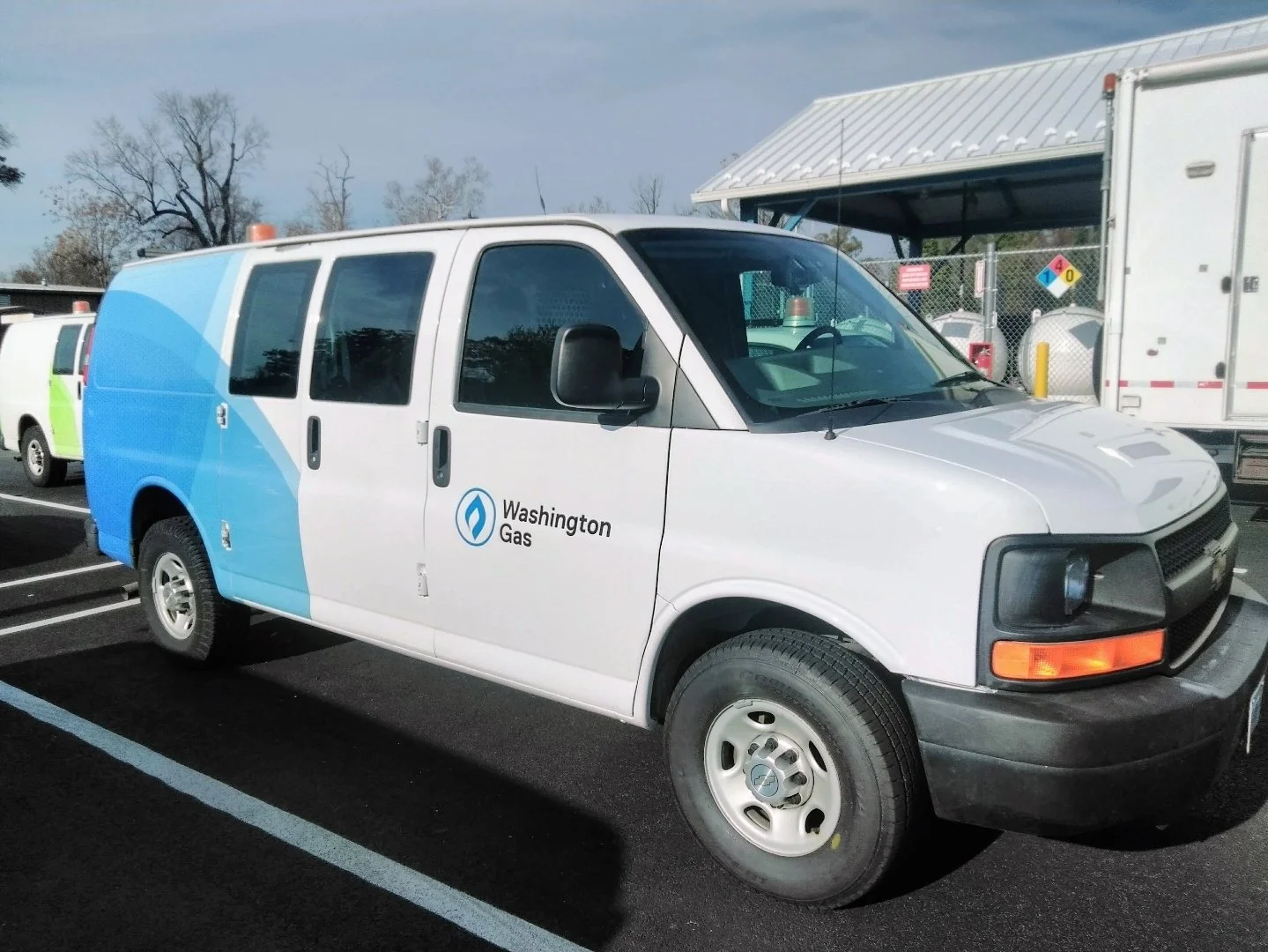 White Washington Gas repair van parked in a lot, with other utility vehicles nearby and a chain-link fence in the background.