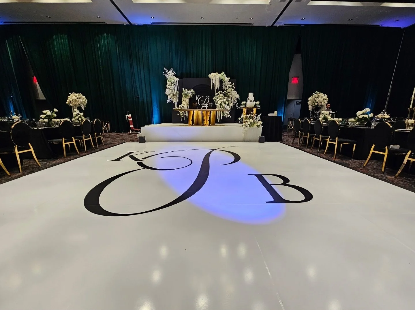 Elegant wedding reception setup with a large dance floor featuring black calligraphy initials, floral arrangements on tables, and a decorated cake table in the background.