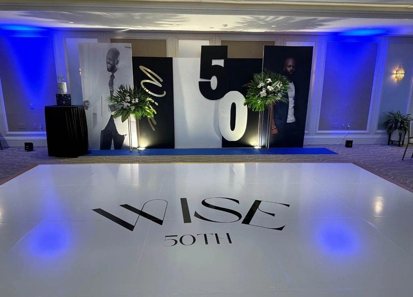 Party setup with large black and white backdrop displaying '50' and portraits of a man on either side, floral arrangements, and a dance floor with 'LISA 50TH' written on it.