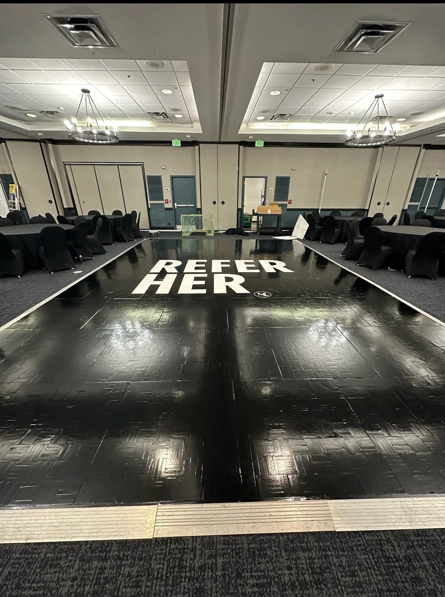 Empty event space with a black dance floor featuring white text that says 'REFER HER'. Surrounding tables with black chairs, and a few items and equipment at the back of the room.