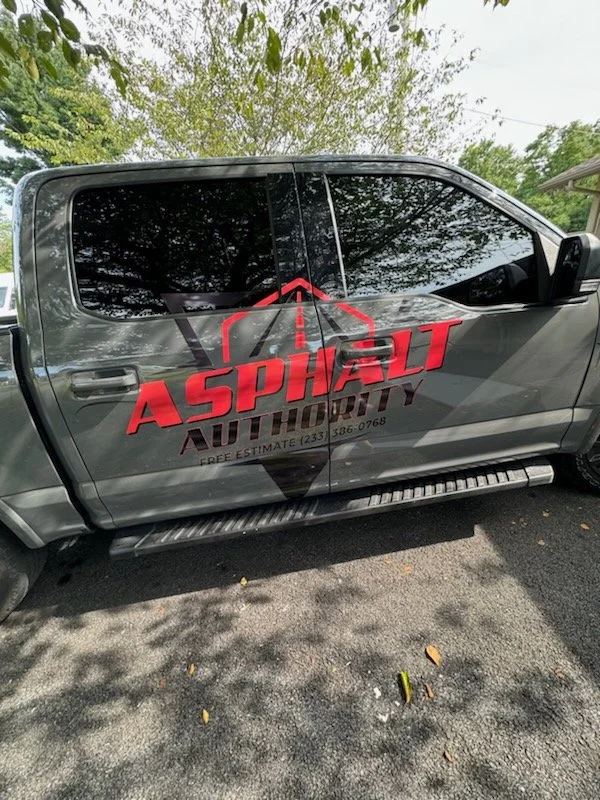 A gray pickup truck displaying a logo for 'Asphalt Authority' on the side, with a large red and black design, parked on a tree-lined street or parking lot.