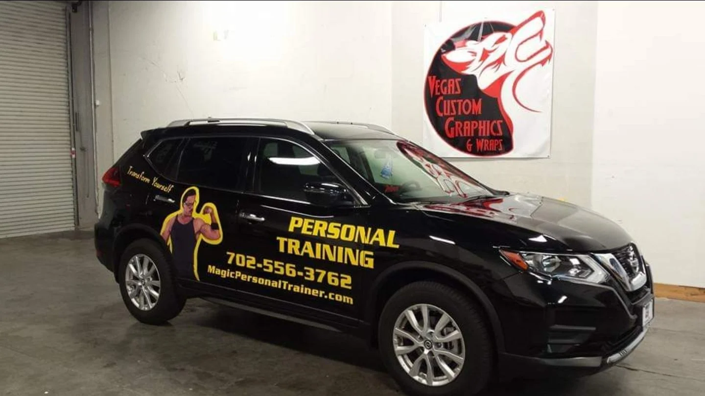 Black SUV with yellow lettering advertising personal training, including a phone number and website, parked indoors near a white wall with a red and black sign that says Vegas Custom Graphics & Wraps.