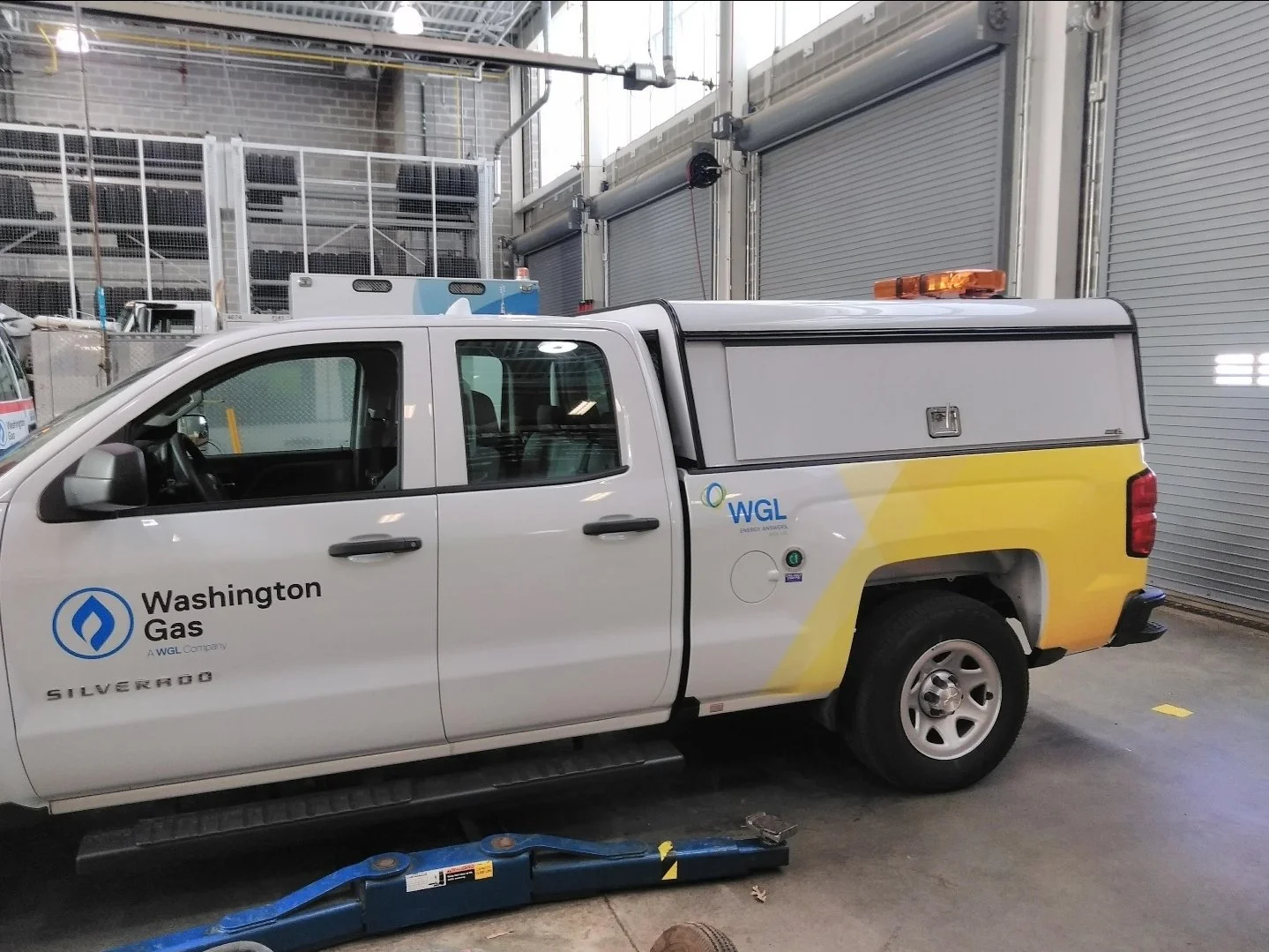 White Washington Gas utility truck parked indoors next to a gray roller door, with a blue hydraulic floor jack beneath it.