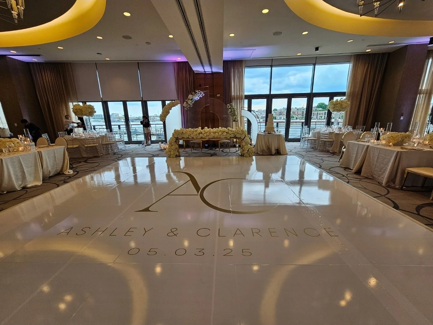 Elegant wedding reception setup with a dance floor in the center, decorated with the couple's names Ashley & Clarence and the date 05.03.25. The room features large windows with city and water views, round tables with white tablecloths and floral cen