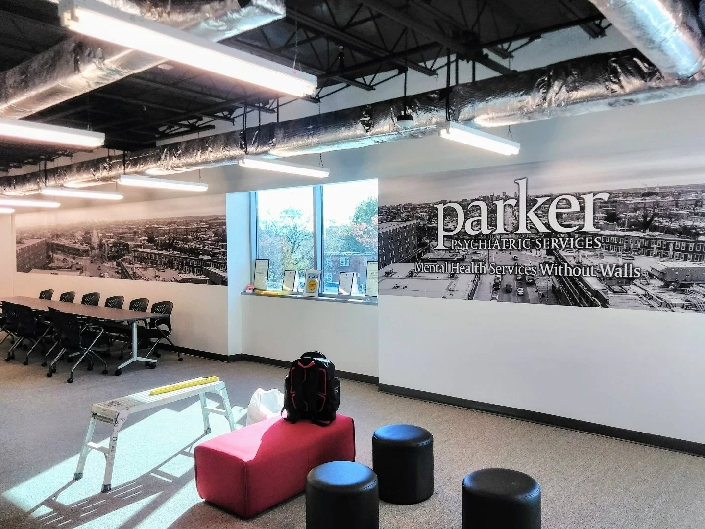 Interior of a psychiatric office with a large special black-and-white cityscape mural wall displaying the words 'Parker Psychiatric Services' and 'Mental Health Services Without Walls.' There are tables, chairs, a window with certificates or awards o