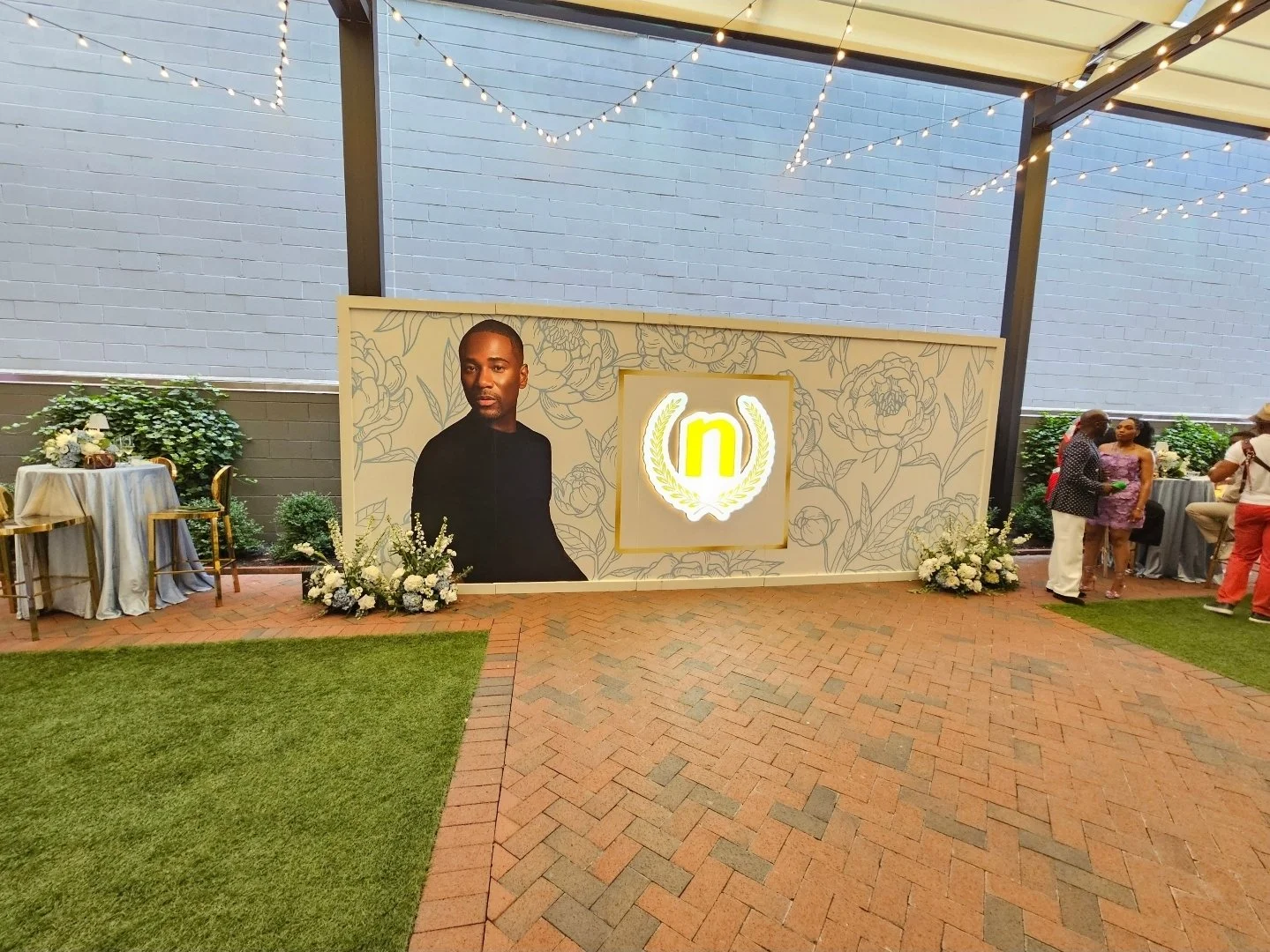 Indoor event space with a large backdrop displaying a photo of a man with a dark shirt, floral patterns, and a bright yellow 'n' logo in a wreath design. There are tables decorated with flowers and guests mingling to the right.