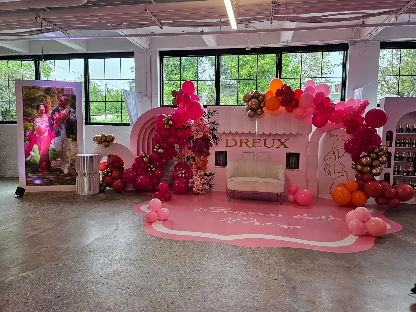 Decorative pink and purple balloon arrangement with flowers in front of a white backdrop with the word Dreux in gold, pink carpet with cursive writing, and a small white bench. Large windows showing green trees outside.