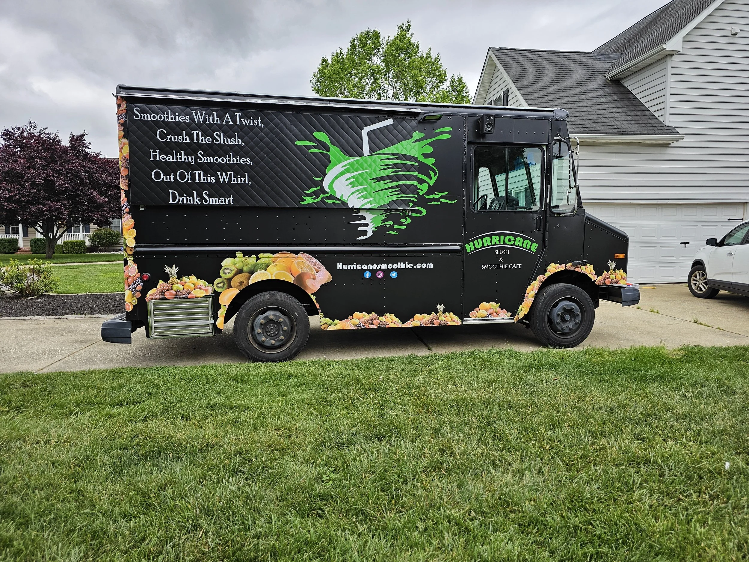 A black food truck decorated with colorful images of fruits, featuring the name Hurricane Smoothie Cafe and advertising smoothies and slush drinks.