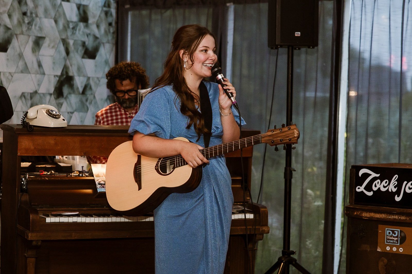 Vrouw met lang haar in een blauwe jurk speelt gitaar en zingt in een kamer met een piano en een man achter de piano, die een retro telefoon vasthoudt.