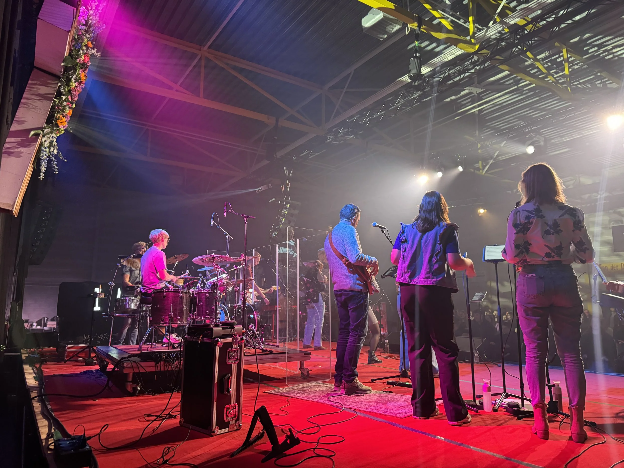 Muziekband op een podium met instrumenten en microfoons, onder kleurrijke podiumlichten en een industrieel plafond.