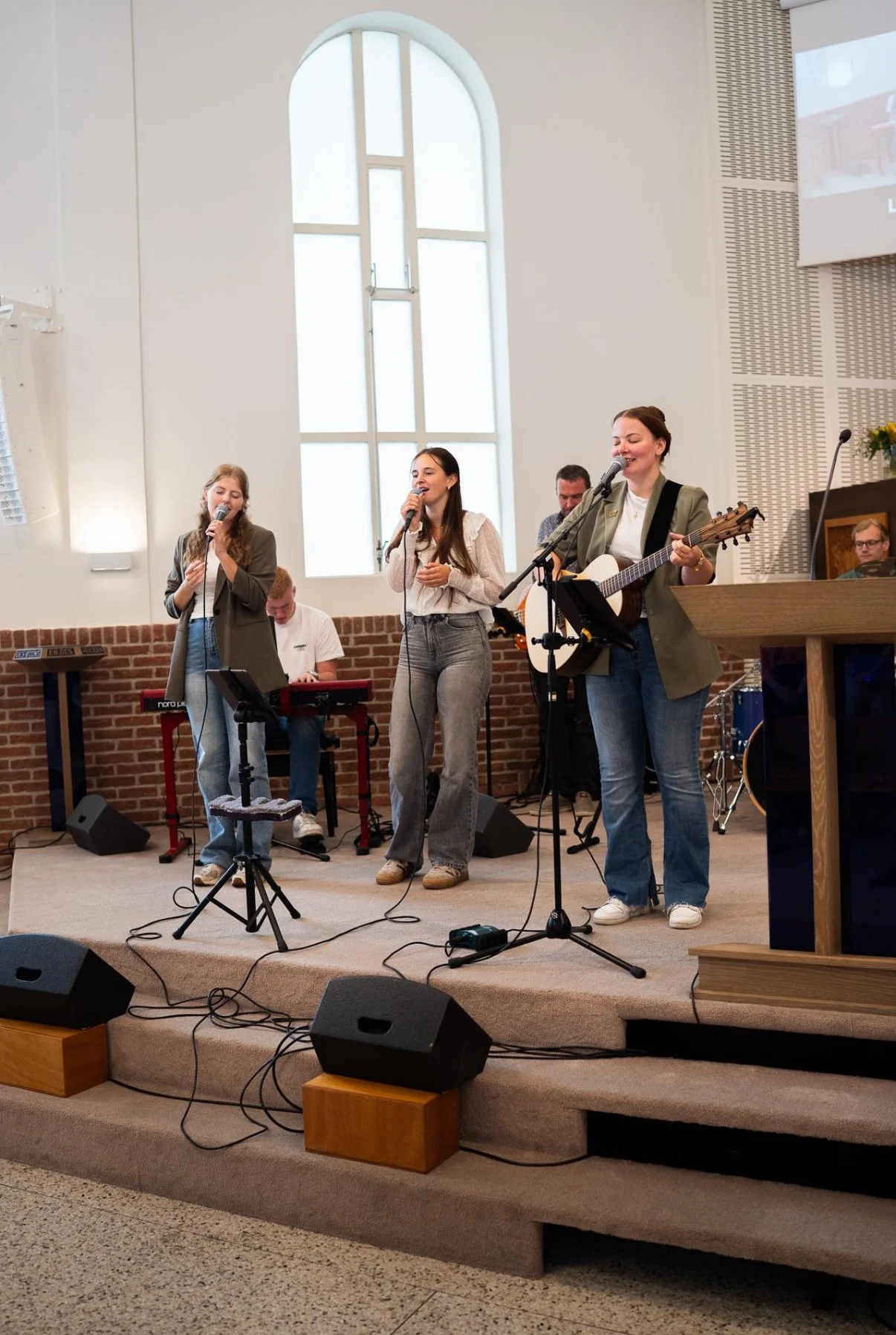 Vrouw met akoestische gitaar en twee zangeressen op een podium in een kerk, met een keyboardspeler en een drummer op de achtergrond.