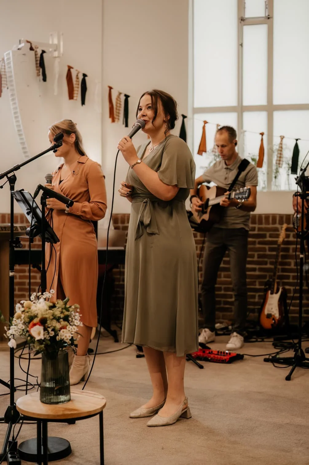 Een band bestaande uit een vrouw die zingt met een microfoon, een vrouw die op een tablet speelt en een man die gitaar speelt, voert live muziek op in een gezellige, binnenruimte met grote ramen en decoraties.