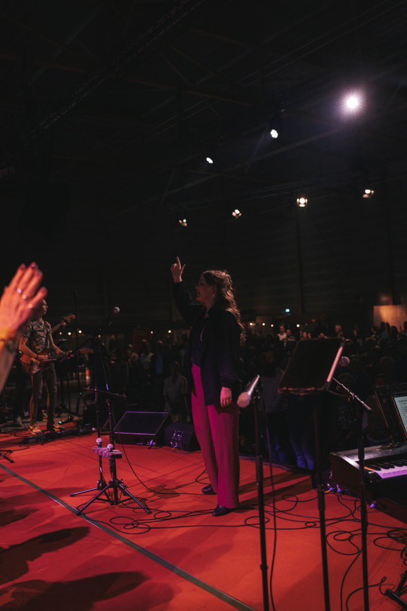 Een vrouw staat op een podium met een microfoon, omringd door muziekapparatuur en een publiek in een donkere concertzaal.