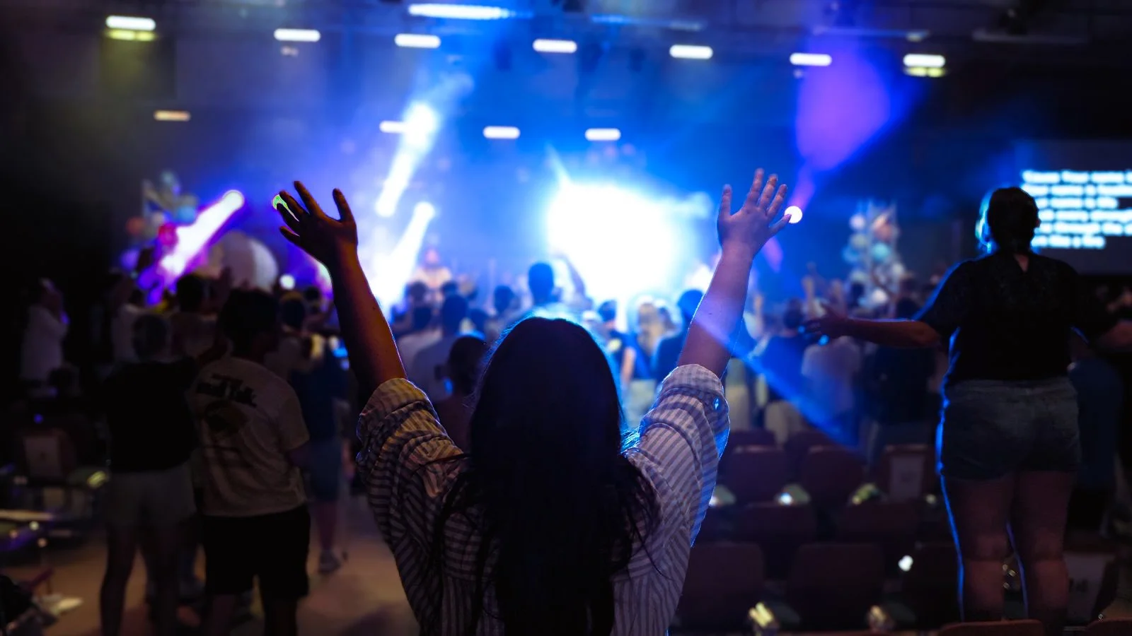 Concert met een vrouw met omhoog gestoken handen in de voorgrond en een volle menigte, blauwe en paarse lichten op het podium.