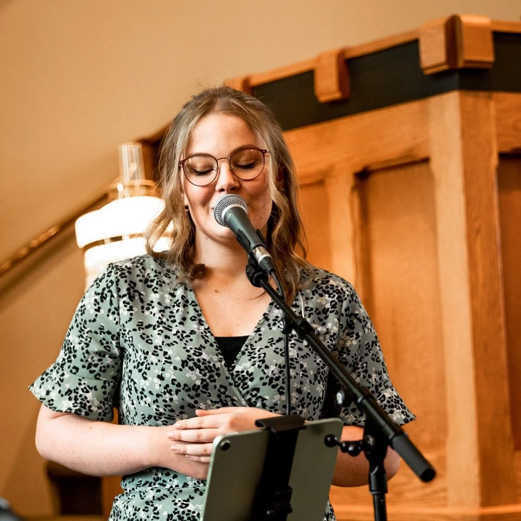 Een vrouw met bril die zingt of spreekt in een microfoon, staand voor een houten decor of meubel, binnen in een kamer met warme verlichting.